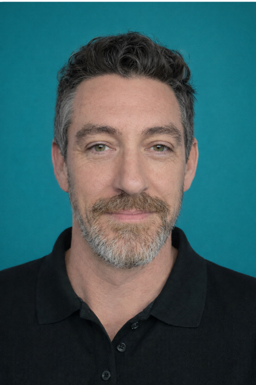 A man with wavy brown hair and a beard is standing in front of a shelf filled with various household items. He is wearing a light blue polo shirt and has a neutral expression.