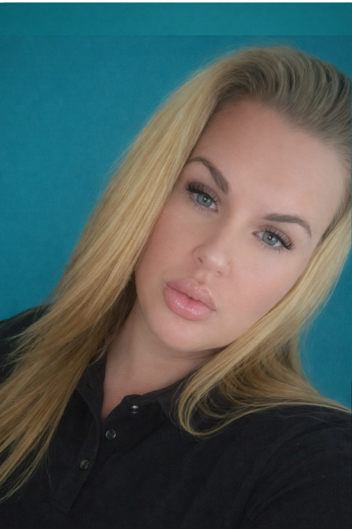 A smiling young woman with shoulder-length dark hair is standing in a bright, modern room with light-colored furniture and minimal decor. She is wearing a plain white t-shirt and blue jeans.