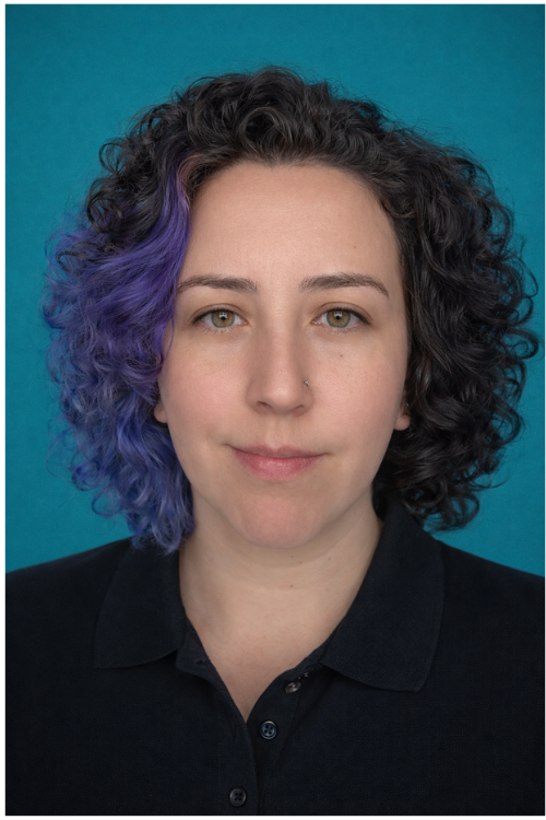 A woman with shoulder-length brown hair, wearing a white blouse, smiles at the camera in a bright room with turquoise and white decorative items on shelves behind her.